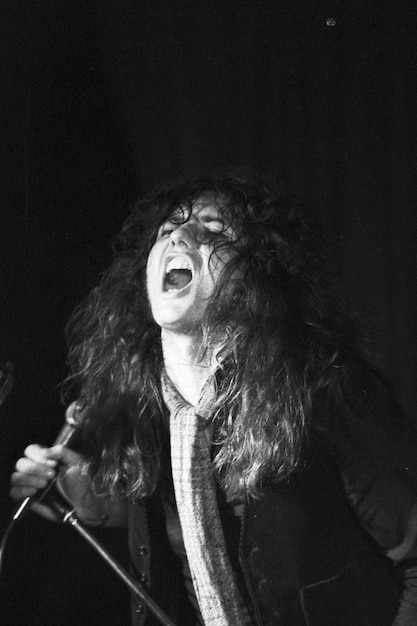 A black and white photo of Tom Araya screaming into a microphone during a concert. The image is intense and captures the energy of his vocal performance. The background is blurred, focusing on Araya's expression of raw emotion.