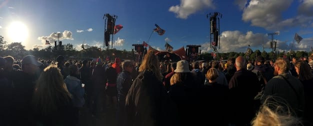A panoramic shot of a diverse crowd at a System of a Down concert, with fans of all ages and backgrounds singing along to the lyrics. The stage is visible in the background, with the band members performing energetically. The overall atmosphere is one of excitement and unity.
