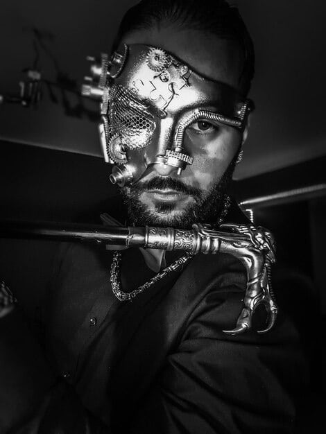 A close-up, high-contrast image of a black metal musician's face, heavily made up with corpse paint, staring intensely into the camera. The background is blurred and dark, emphasizing the musician's unsettling and theatrical appearance.
