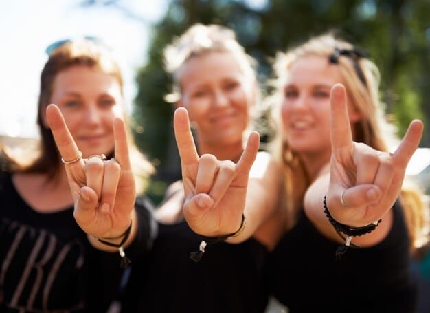 A group of heavy metal fans wearing band t-shirts, posing for a photo together at a concert venue. They are smiling and holding up metal horns, showcasing the camaraderie and shared passion within the fan club community.