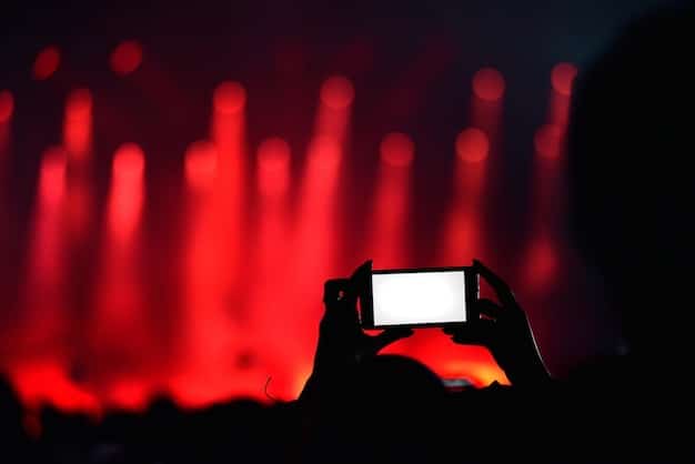 A close-up shot of a person holding a smartphone, discreetly recording a concert in a dark venue. The stage is visible in the background, with bright lights and silhouettes of performers.