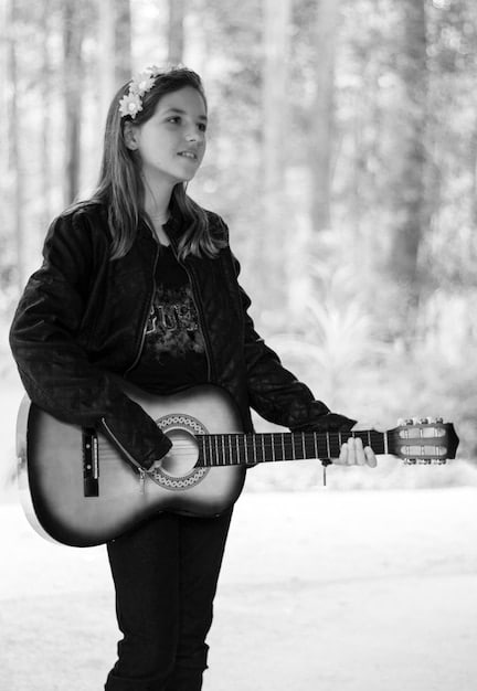A black and white photo of a young aspiring rock musician (around 14 years old) practicing guitar in a cluttered bedroom. Posters of classic rock bands adorn the walls, and the room is filled with musical instruments and amplifiers.